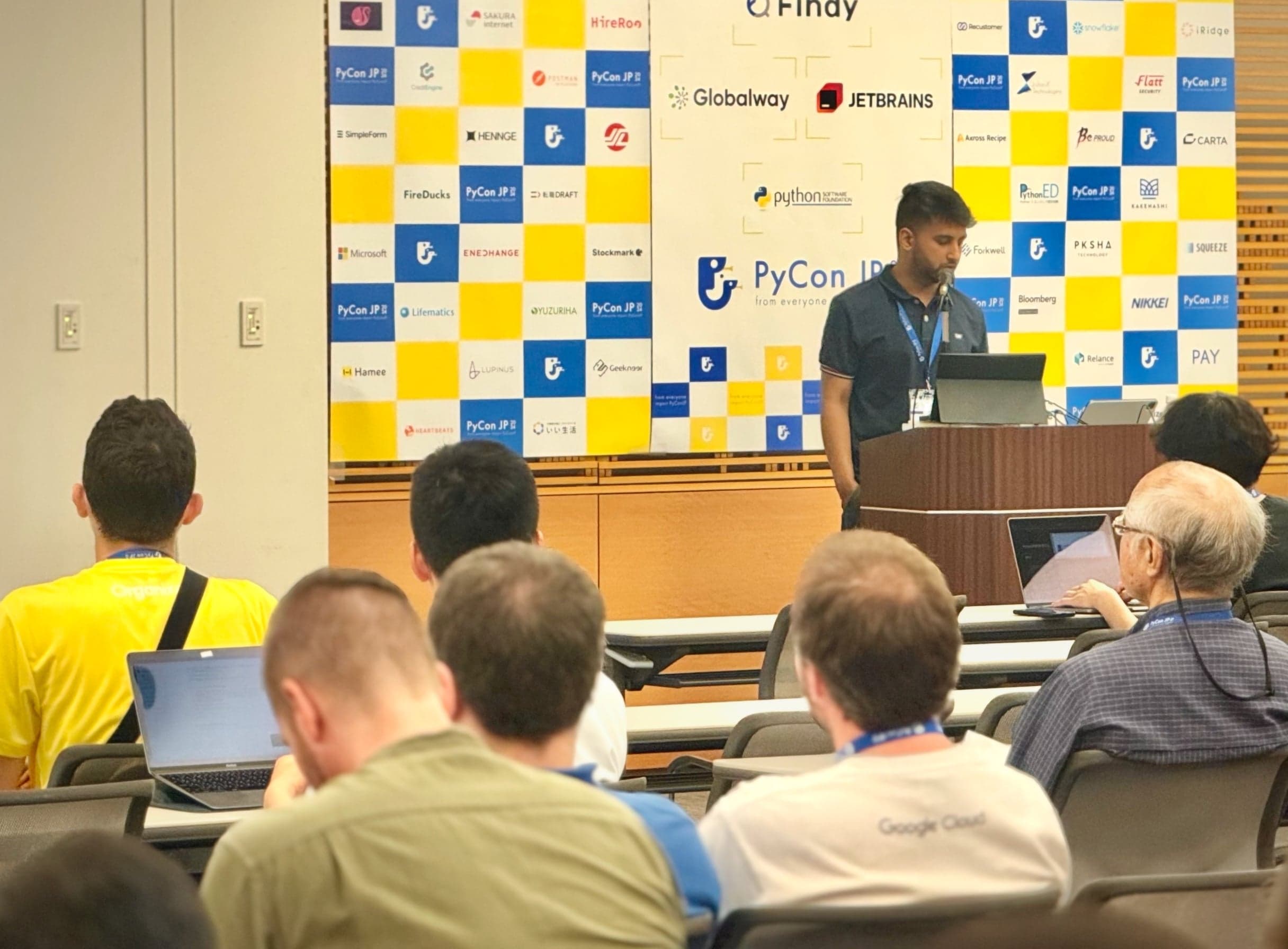 Nirmal presenting from a podium in front of the PyCon JP 2024 sponsor banner to a seated audience